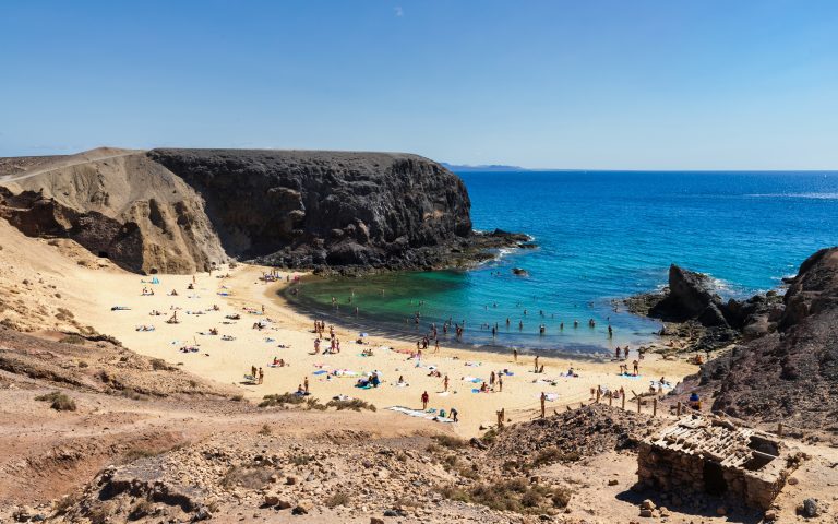 This secluded golden sand beach in Canary Islands is cradled by dramatic, rugged desert cliffs and striking turquoise waters where sunbathers and swimmers enjoy a pristine, sun-drenched cove.